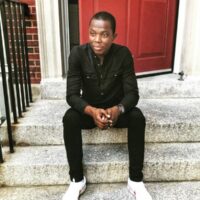 Romeo Oriogun sits outside on the front steps of a building dressed in black with white shoes