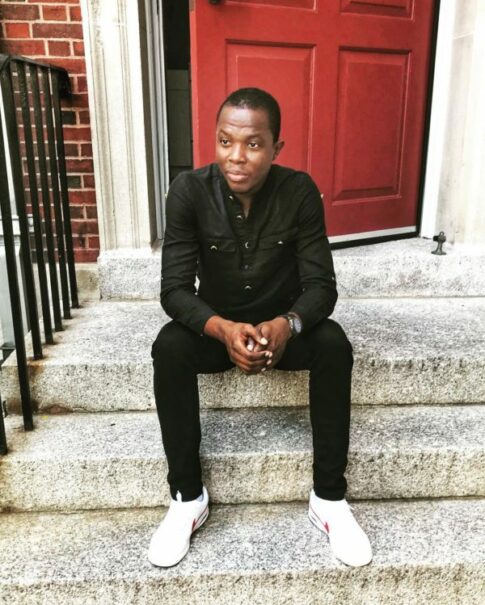 Romeo Oriogun sits outside on the front steps of a building dressed in black with white shoes